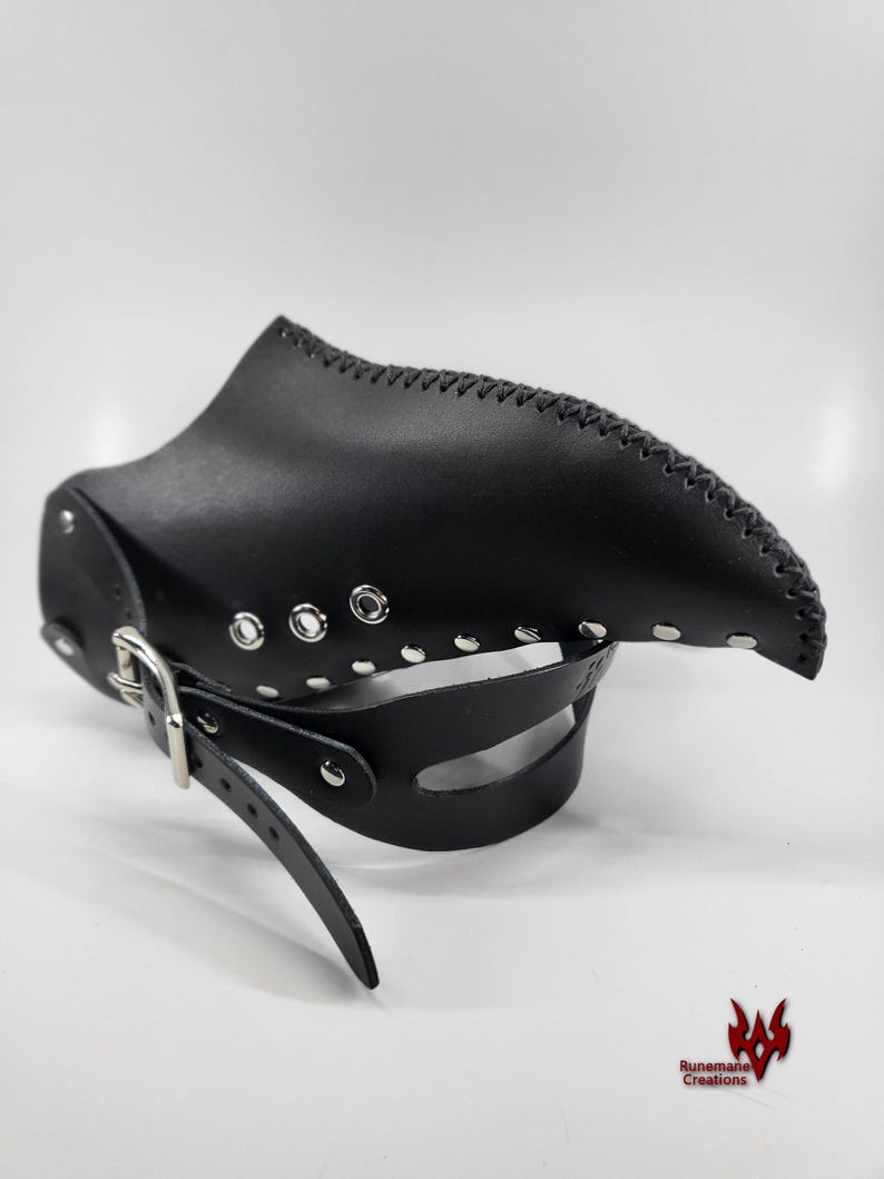 Puede incluir: Una m&aacute;scara de cuero negro con un dise&ntilde;o puntiagudo y curvo. La m&aacute;scara tiene una correa con una hebilla y m&uacute;ltiples ojales y tachuelas plateadas.