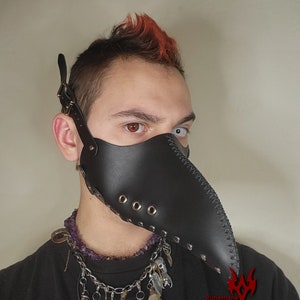 Puede incluir: Una m&aacute;scara de m&eacute;dico de la peste de cuero negro con un pico largo y remaches met&aacute;licos. La m&aacute;scara la lleva una persona con el pelo rojo y un collar con abalorios.