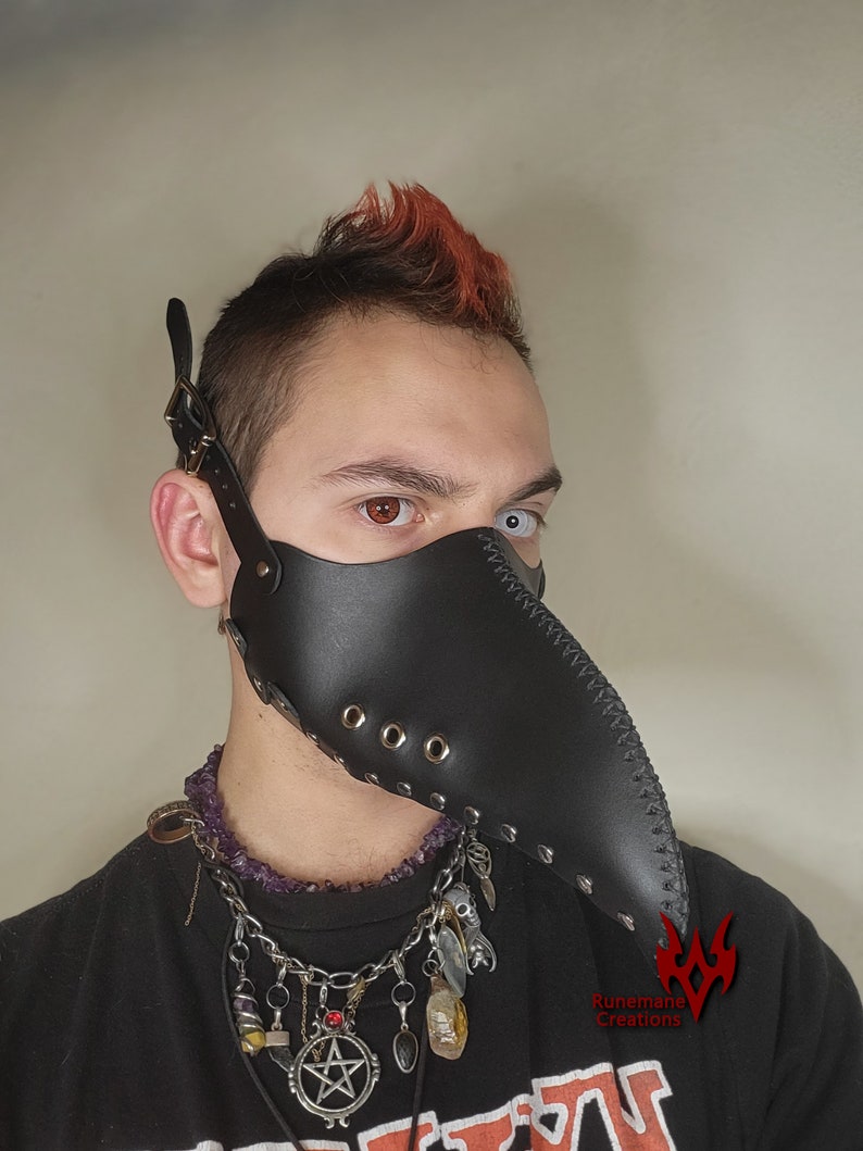 Puede incluir: Una m&aacute;scara de m&eacute;dico de la peste de cuero negro con un pico largo y remaches met&aacute;licos. La m&aacute;scara la lleva una persona con el pelo rojo y un collar con abalorios.