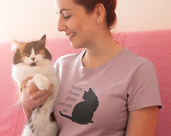 May include: A light purple short-sleeved t-shirt featuring a black graphic of a sitting cat and the text "Sorry, I have plans with my cat." The shirt has a crew neckline and is made of a solid-colored fabric with a simple, casual design.