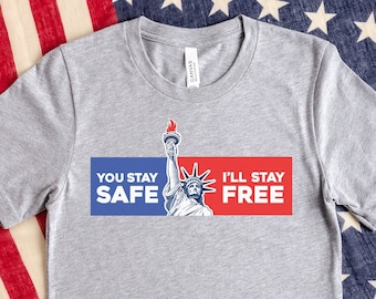 Puede incluir: Una camiseta gris jaspeado de manga corta con un gráfico de la Estatua de la Libertad entre un rectángulo azul con el texto blanco YOU STAY SAFE y un rectángulo rojo con el texto blanco I'LL STAY FREE, con una llama de antorcha roja.