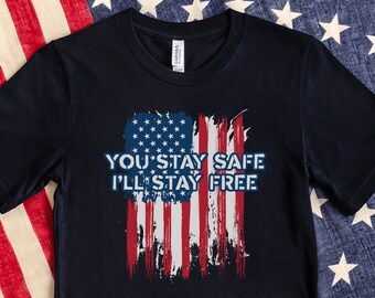 Puede incluir: Una camiseta negra de manga corta con un gráfico de la bandera estadounidense desgastada en el centro. El texto "YOU STAY SAFE I'LL STAY FREE" está impreso en letras mayúsculas blancas sobre el diseño de la bandera, que tiene rayas rojas y blancas.