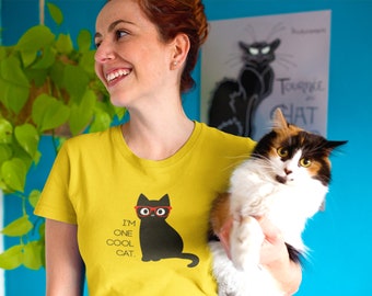 May include: A mustard yellow short-sleeved t-shirt featuring a graphic of a black cat wearing red glasses. The text printed below the cat reads, I'M ONE COOL CAT. The shirt has a crew neckline and is made of a solid-colored cotton fabric.