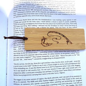 May include: A wooden bookmark with a fishing theme. The bookmark features an etched design of a fish, a fishing rod, and a line. A dark brown cord is attached for easy placement in a book.