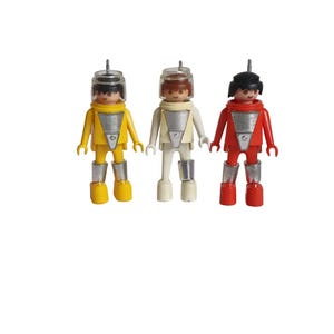 May include: Three Playmobil astronaut figures in yellow, white, and red space suits. Each figure has a clear helmet, silver boots, and a small antenna. The figures are standing upright against a white background.