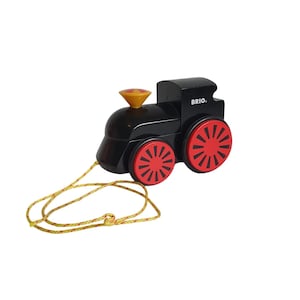 May include: Black wooden toy train with red wheels and a yellow rope. The train has a yellow and orange chimney and the word "BRIO" printed on the side. A classic pull-along toy.