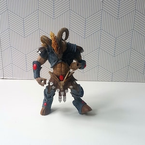 May include: A blue and brown plastic action figure of a horned warrior with red accents. The figure is standing on a white surface.