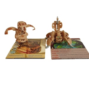 Op de afbeelding: Twee bruine plastic speelgoedfiguren met kaarten. De figuren zijn van de Bakugan Battle Brawlers speelgoedlijn. De kaarten hebben de namen "Driger" en "Wilda" erop gedrukt.