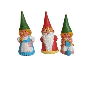 Könnte beinhalten: Drei kleine Gnomfiguren. Ein weiblicher Gnom in einem blauen Kleid und grüner Mütze, ein männlicher Gnom in einem roten Gewand und grüner Mütze und ein weiblicher Gnom in einem rosa Kleid und grüner Mütze.
