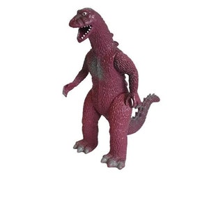 Può includere: Un dinosauro di plastica rosso giocattolo con una superficie testurizzata e una bocca spalancata. Il dinosauro ha una lunga coda e sta in piedi su uno sfondo bianco.