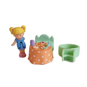 Puede incluir: Una muñeca en miniatura con coletas rubias está de pie cerca de una pequeña mesa preparada para una comida. La mesa está cubierta con un mantel naranja con lunares. También hay una silla verde y una pequeña bañera verde.