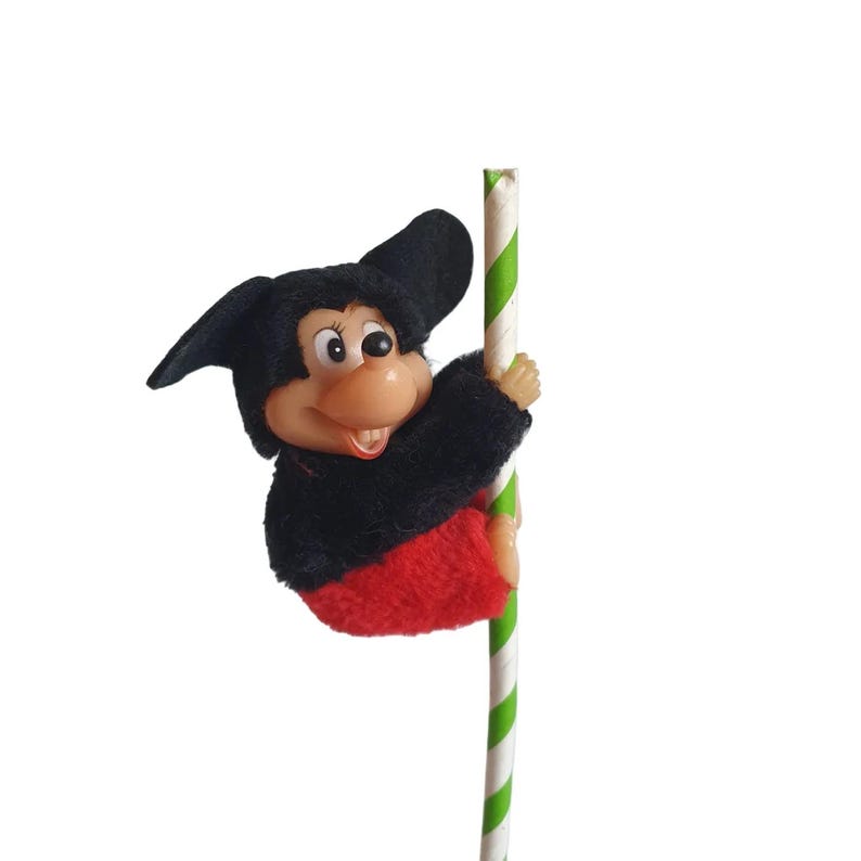 May include: A vintage Mickey Mouse toy figure, dressed in black and red, clinging to a green and white striped pole. The figure has a smiling face and large black ears. The toy is set against a white background.
