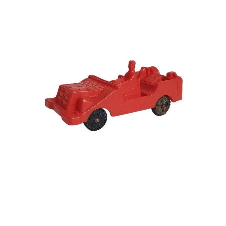 May include: A small, red toy jeep with black wheels and a driver figure. The vintage vehicle has a simple design, with a flat front, open top, and a rear cargo area. The wheels are black and the jeep is set against a white background.