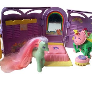 以下が含まれることがあります： ピンクのベッド、緑とピンクのグルーミングステーション、ピンクのたてがみと尻尾を持つ薄い緑のポニーが付いた、紫と黄色のプラスチック製のポニー用プレイハウスです。