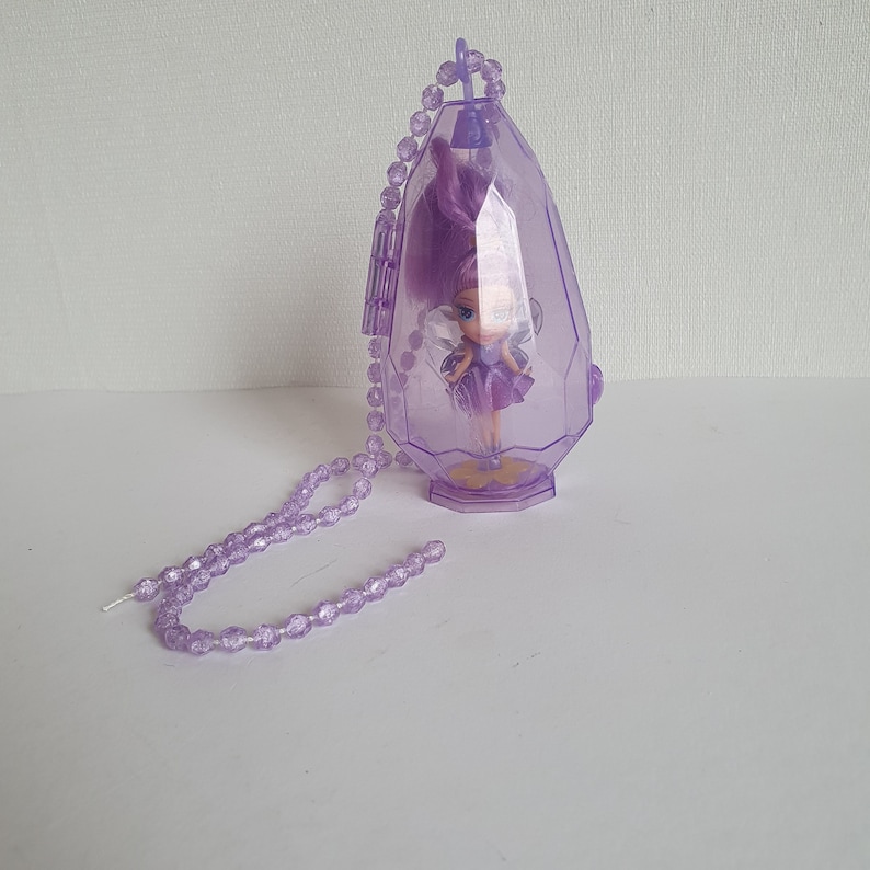 May include: A purple plastic necklace with a clear pendant shaped like a gem. Inside the pendant is a small purple fairy doll with wings.