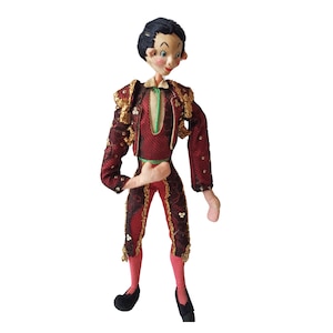 May include: A vintage doll dressed as a matador. The doll has black hair, a red and gold costume, and a green scarf.