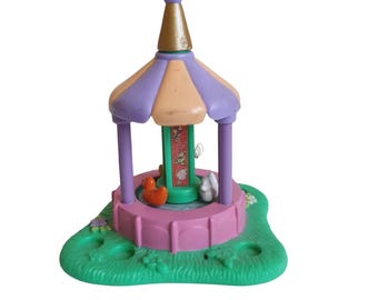 May include: A vintage toy carousel with a purple and peach striped canopy, supported by purple columns. The base is pink, with a green grassy area. Inside, there's a duck and a rabbit. The carousel has a gold-colored top.