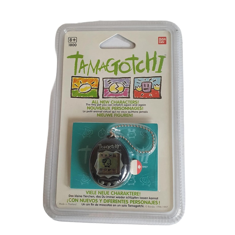 May include: A black and gray Tamagotchi keychain toy in its original packaging. The packaging is white with green and red accents. The text on the packaging reads "Tamagotchi" and "All New Characters!"