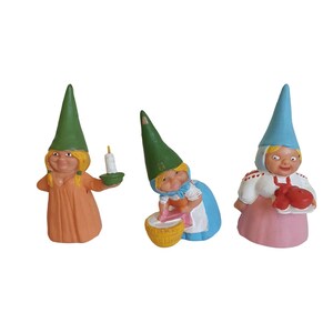 May include: Three small ceramic figurines of gnomes with pointed hats. The gnome on the left is wearing a green hat and an orange dress and holding a candle. The gnome in the middle is wearing a green hat and a blue dress and holding a small basket. The gnome on the right is wearing a blue hat and a pink dress and holding two red apples.