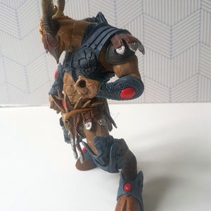 May include: A brown and gray action figure of a horned creature wearing armor. The figure has red accents on its armor and is posed in a dynamic stance.