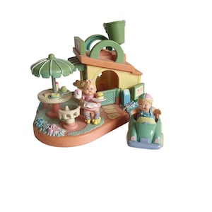 Puede incluir: Un juego de miniaturas que representa una escena de cafetería con dos figuras. La cafetería tiene una sombrilla verde, una mesa con pasteles y un coche pequeño. Las figuras visten colores pastel, y el diseño es caprichoso.