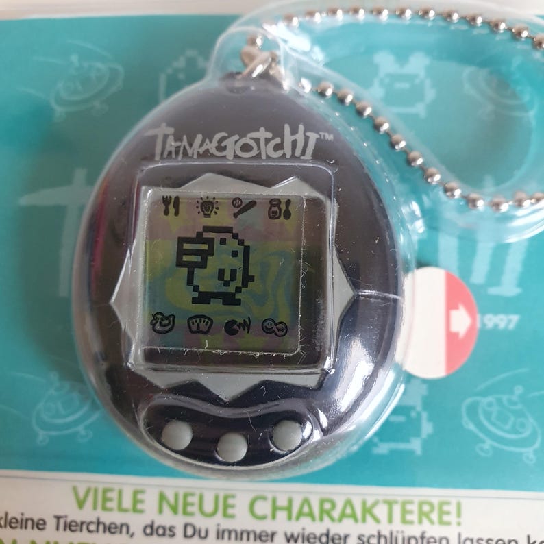 May include: A black Tamagotchi keychain with a digital screen displaying a pixelated character. The screen also shows icons for feeding, playing, and other activities. The text "TAMAGOTCHI" is printed on the front of the device.