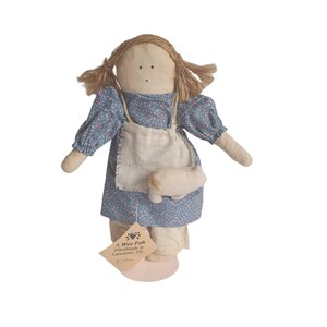 May include: A handmade cloth doll with brown hair, wearing a blue dress with pink flowers and a white apron. The doll is holding a small white stuffed animal and has a tag that reads "A Wee Folk Handmade in Lancaster, PA".