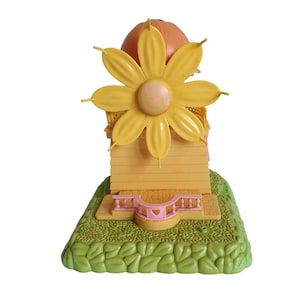 May include: A yellow and orange plastic toy windmill with a flower-shaped top and a pink heart on the base. The windmill is on a green base with a leafy design.