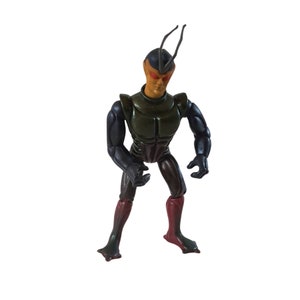 May include: A green and black action figure with a bug-like appearance. The figure has a black head with two long antennae, orange eyes, and a green and black body. The figure is standing with its arms bent at the elbows and its fists clenched.