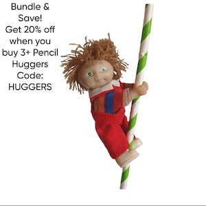 Peut inclure: Une petite poupée aux cheveux bruns et aux yeux verts, vêtue d'une tenue rouge, s'accroche à un poteau rayé vert et blanc. La poupée est un Pencil Hugger. Le texte de l'image indique : Bundle & Save ! Obtenez 20 % de réduction à l'achat de 3+ Pencil Huggers. Code : HUGGERS.
