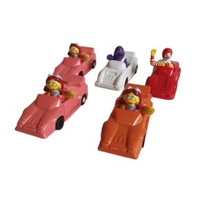 May include: A collection of vintage McDonald's Happy Meal toys featuring various characters in race cars. The cars are in shades of pink, red, orange, and white. Each car has the McDonald's logo.