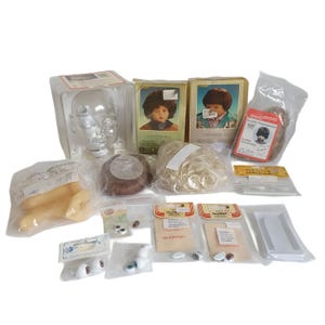 Op de afbeelding: Een verzameling poppenmaakbenodigdheden, waaronder poppenpruiken, ogen en een doorzichtige plastic poppenkop. De items zijn verpakt in doorzichtige plastic zakken en dozen. De pruiken zijn in bruine en blonde tinten.