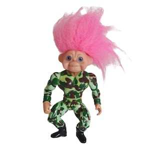May include: A pink haired troll doll wearing a green and brown camouflage outfit. The doll has blue eyes and a friendly expression.