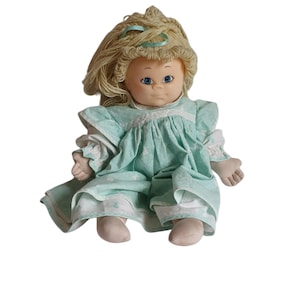 Może przedstawiać: Lalka vintage z blond włosami z włóczki, niebieskimi oczami i jasnoniebieską sukienką z białą lamówką. Lalka siedzi ze skrzyżowanymi nogami.