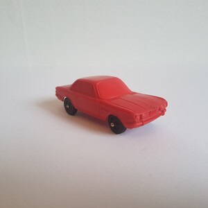 May include: A red toy car with black wheels. The car is a coupe style with a rounded front end and a long hood.