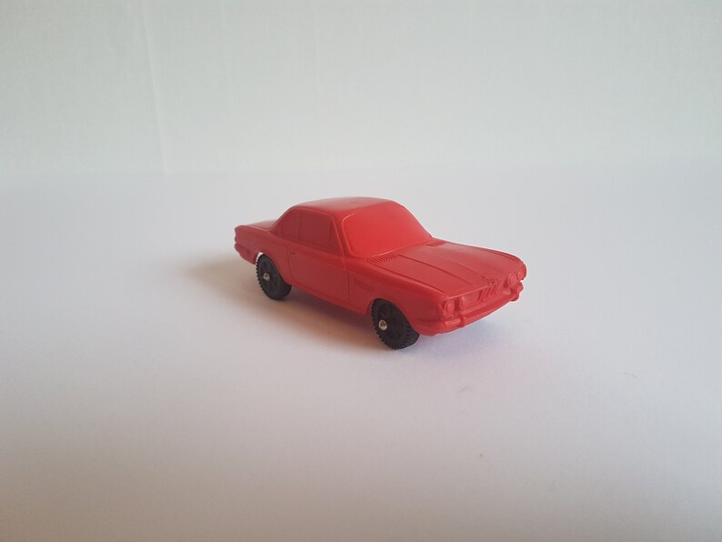 May include: A red toy car with black wheels. The car is a coupe style with a rounded front end and a long hood.