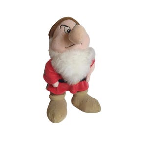 Könnte beinhalten: Ein Plüschtier von Grumpy, einem der sieben Zwerge aus dem Disney-Film Schneewittchen und die sieben Zwerge. Grumpy trägt ein rotes und schwarzes Outfit mit einem weißen Bart und hat einen mürrischen Gesichtsausdruck.