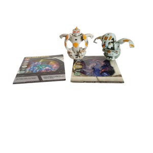 May include: Two white and orange Bakugan toys with their corresponding cards. The cards feature the Bakugan's name, "Colorstorm", and stats. The cards are white with black and blue text and images.