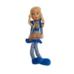 Puede incluir: Una muñeca de trapo vintage con trenzas rubias, ojos azules y labios rojos. La muñeca viste un vestido con estampado floral con un delantal azul y una falda con volantes azules. Las largas piernas de la muñeca son a rayas azules y blancas, con pies azules.