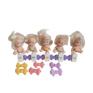 Puede incluir: Cinco pequeñas muñecas bebé con cabello rubio, que visten pañales de diferentes colores. Están sentadas frente a una valla púrpura con detalles en forma de corazón y pequeños objetos con forma de cartón de leche. Debajo hay varios accesorios para el cabello en forma de lazo.