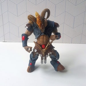 May include: A brown and blue action figure of a horned creature with a red gem on its chest. The figure is wearing a leather skirt and armor.