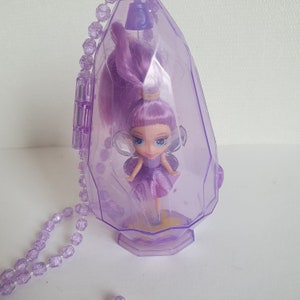 May include: A purple plastic necklace with a clear plastic pendant containing a small purple fairy doll with wings. The doll has long purple hair and a purple dress.