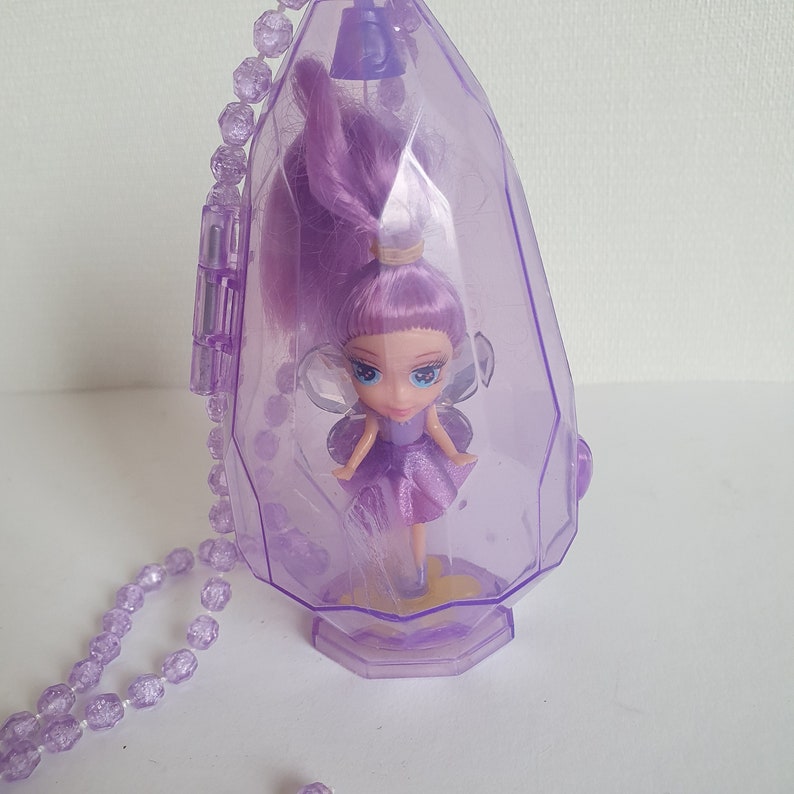 May include: A purple plastic necklace with a clear plastic pendant containing a small purple fairy doll with wings. The doll has long purple hair and a purple dress.