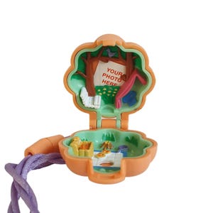 May include: An open, orange, flower-shaped toy with a purple cord. The interior features a miniature scene with trees, a swing, and the text "YOUR PHOTO HERE". The toy is designed to open and close.