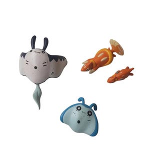 May include: A collection of four cartoon-style sea creature figurines. The figurines include a white and black manta ray, a light blue manta ray, and two orange and yellow shrimp-like creatures. The figurines have cartoon faces.