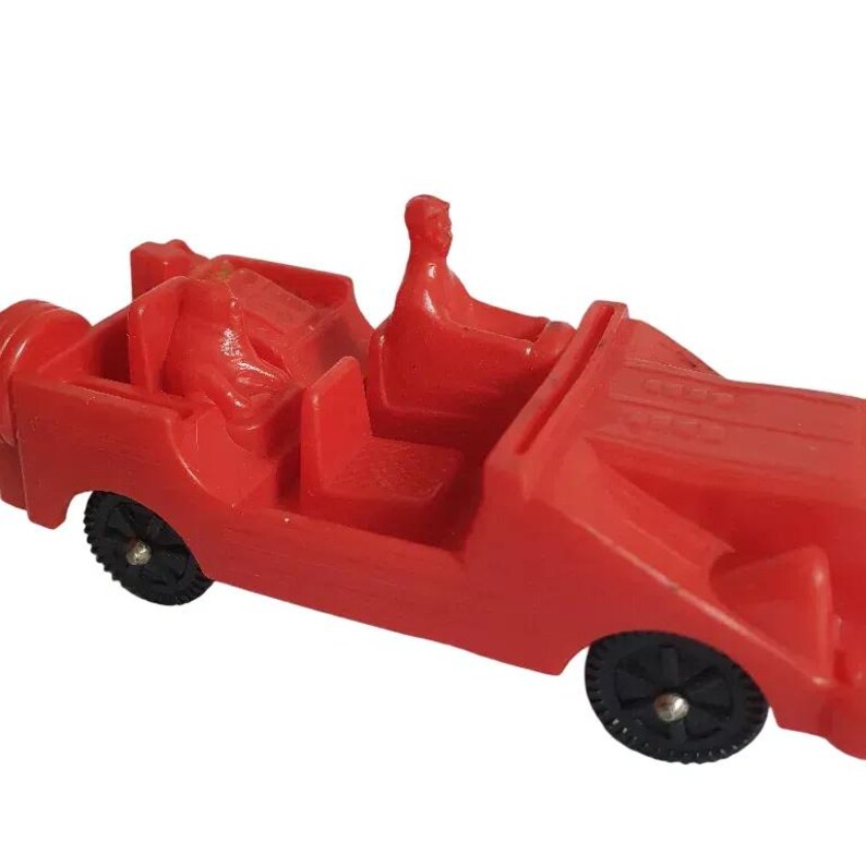May include: A bright red toy car with black wheels. The car has a driver figure and two passenger seats. The toy car is a vintage style, likely from the mid-20th century.