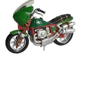 Pode incluir: Uma motocicleta de brinquedo com carenagem verde, para-brisa transparente e assento preto. O chassi apresenta detalhes em vermelho e prata, incluindo um bloco de motor prateado e escapamento duplo. As rodas são pretas com raios prateados.