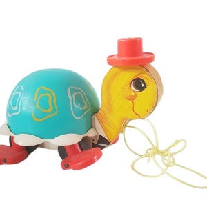 Op de afbeelding: Een vintage trek-speelgoed schildpad met een geel hoofd, een blauwe schild en een rode hoed. De schildpad heeft een gele touw aan zijn hoofd.