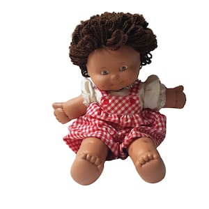 Może przedstawiać: Lalka z brązowymi włosami ubrana w czerwoną i białą kratę w sukienkę z białym gorsetem i czerwonym guzikiem. Lalka ma brązowe oczy i jasnobrązową cerę.