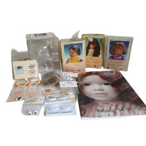 Op de afbeelding: Een verzameling poppenmaakbenodigdheden, waaronder poppenhoofden, pruiken, ogen en een catalogus. De catalogusomslag toont een pop met blauwe ogen en rood haar. Diverse verpakte items zijn zichtbaar, samen met een doorzichtig poppenhoofd.